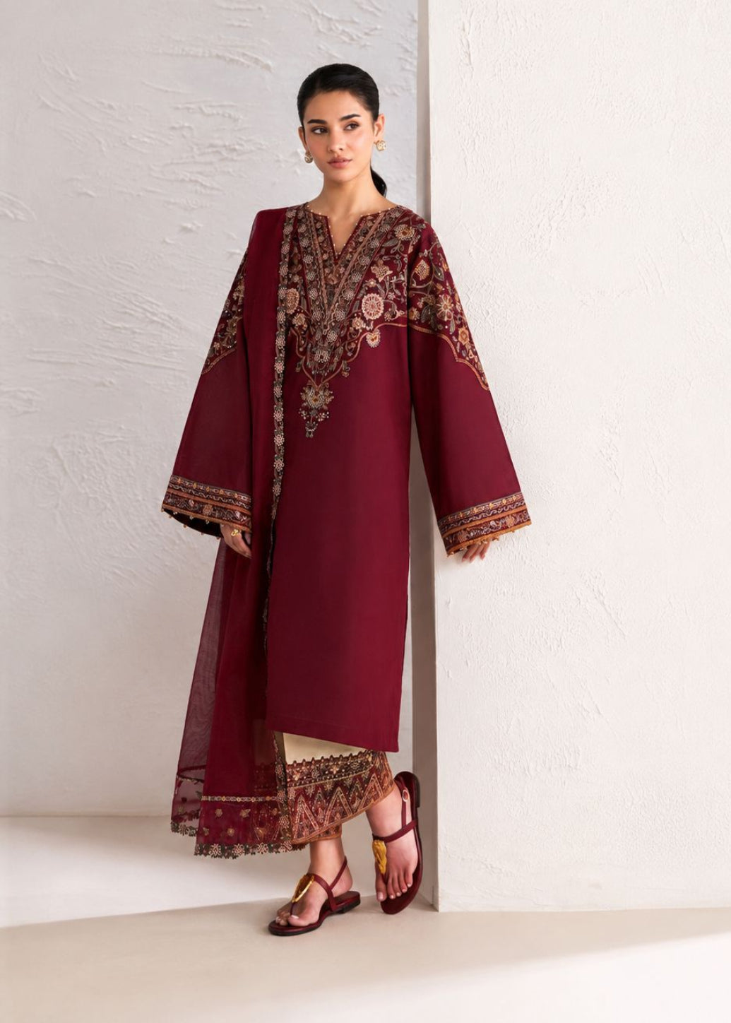 A woman models a traditional deep maroon three-piece kurta set with intricate floral embroidery and border detailing, consisting of a long kurta, matching trousers, and a dupatta, set against a minimalist off-white textured wall.