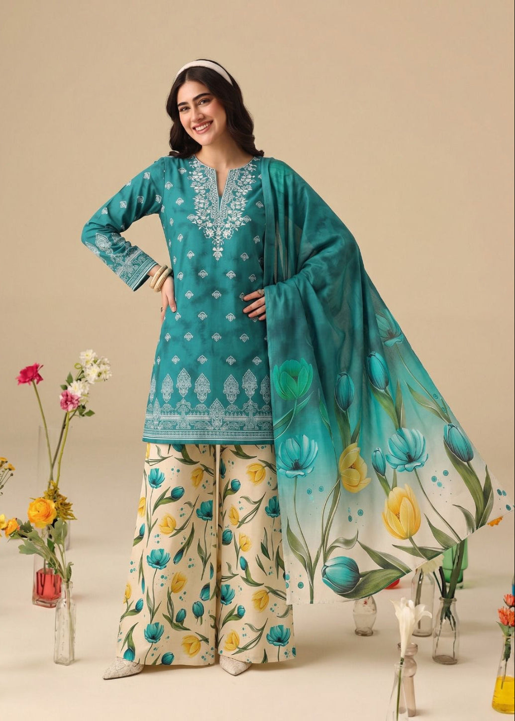 A woman models a traditional South Asian salwar kameez suit set. The set features a teal green kurta with white embroidery, beige wide-leg trousers with a yellow and blue floral print, and a matching teal dupatta with a blue and yellow tulip pattern.