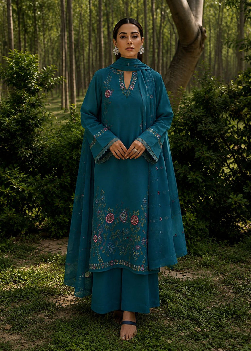 A woman stands outdoors in a wooded setting wearing a teal traditional outfit with floral embroidery and a matching dupatta.
