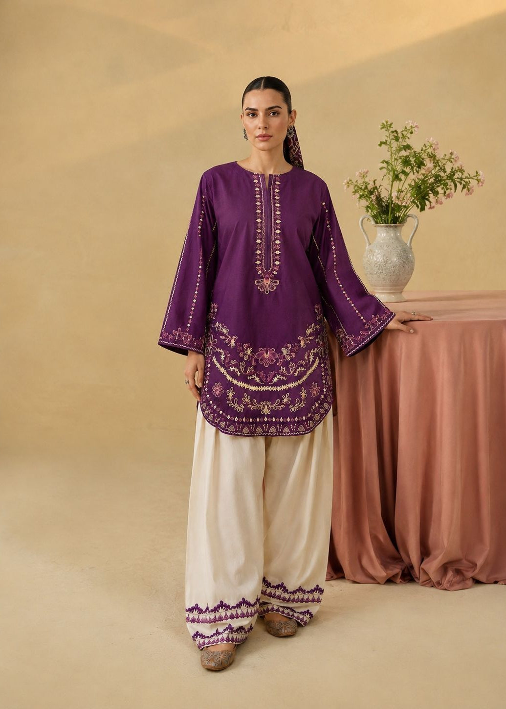 A woman models a traditional South Asian outfit consisting of a purple embroidered kurta and off-white wide-leg palazzo pants with purple borders, standing in a studio with a beige background and a draped table with flowers.
