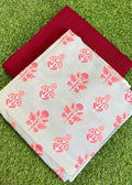 A folded light gray fabric with a coral-pink floral block print pattern, layered over a solid deep red fabric, both laid flat on a green grass background.