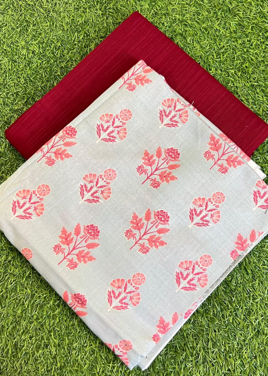 A folded light gray fabric with a coral-pink floral block print pattern, layered over a solid deep red fabric, both laid flat on a green grass background.