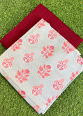 A folded light gray fabric with a coral-pink floral block print pattern, layered over a solid deep red fabric, both laid flat on a green grass background.