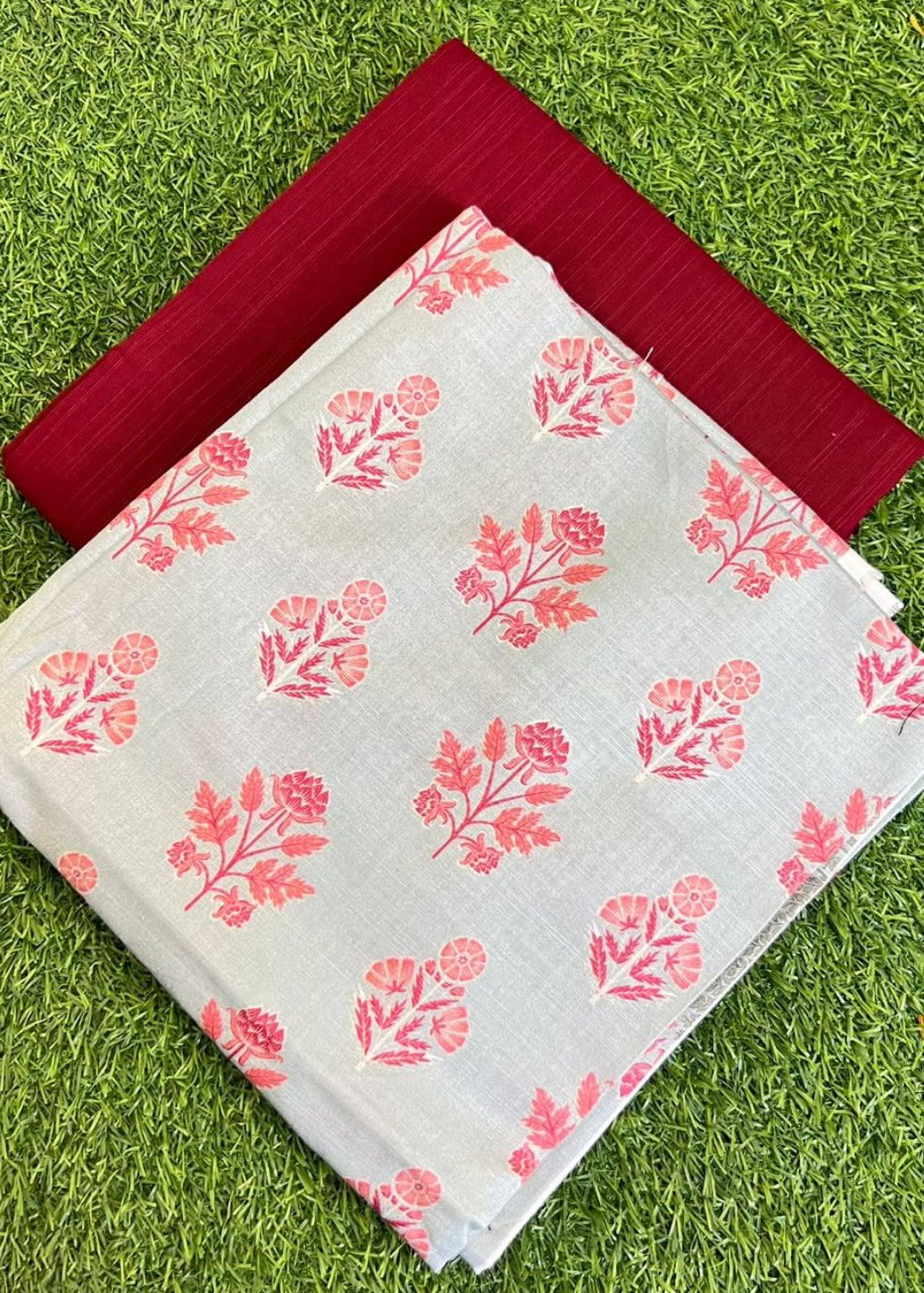 A folded light gray fabric with a coral-pink floral block print pattern, layered over a solid deep red fabric, both laid flat on a green grass background.