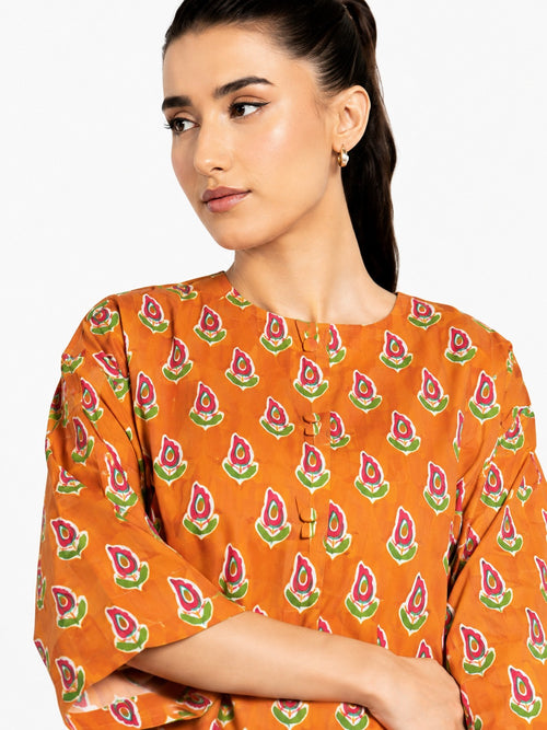 A woman wearing a bright orange kurta with a repeating green and pink block print pattern, shown in a close-up shot against a plain white background.