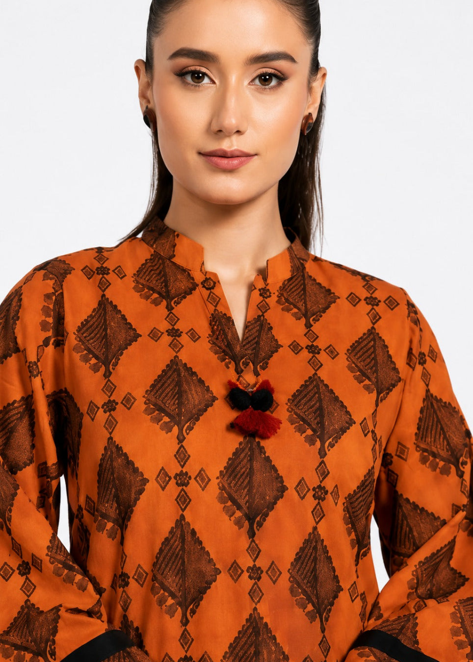 A woman wearing a vibrant orange kurta with a dark brown geometric block print pattern and a decorative black and red tassel tie at the collar, set against a plain light gray background.