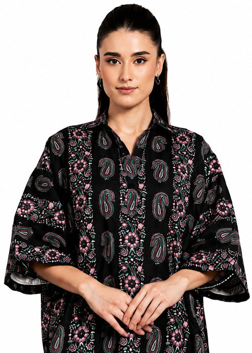 A woman modeling a black kaftan-style top with pink, green, and white paisley and floral patterns, set against a plain white background.