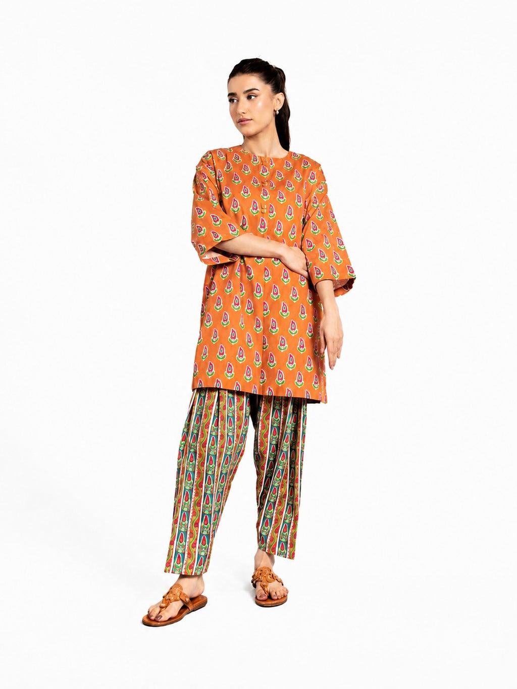 A woman modeling a bright orange kurta with a colorful paisley print and striped palazzo pants, standing against a plain white background.