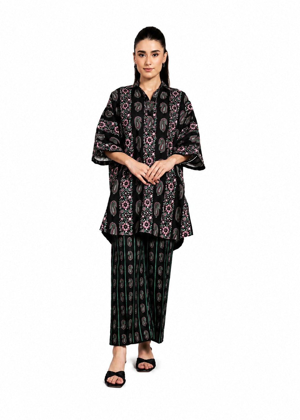 A woman modeling a black two-piece traditional outfit with intricate multicolor floral and paisley patterns, consisting of a short-sleeved tunic and matching wide-leg pants, against a plain white background.