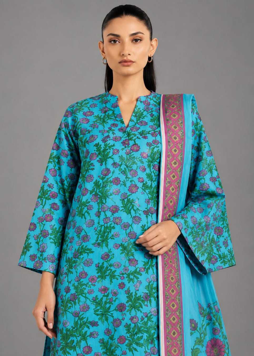 A woman modeling a turquoise blue printed kurta with green and pink floral motifs, paired with matching blue pants and a patterned blue dupatta, against a solid gray studio background.