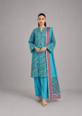 A woman modeling a traditional South Asian outfit consisting of a turquoise blue kurta with green floral patterns, matching turquoise pants, and a turquoise dupatta with a purple and pink border, set against a solid gray studio background.