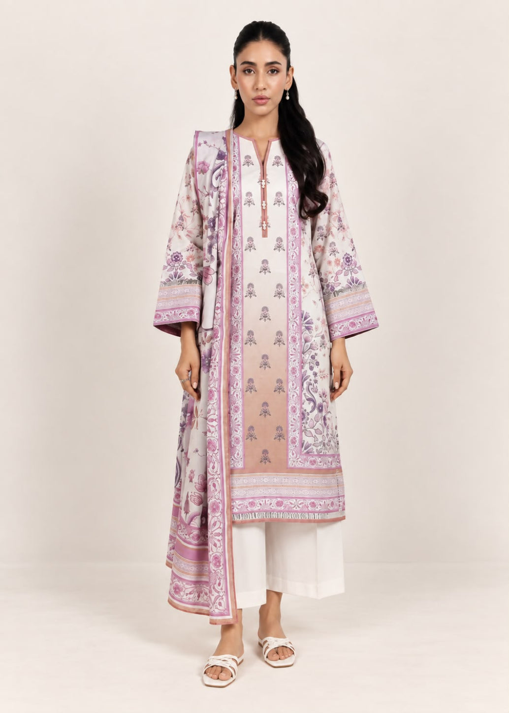 A woman modeling a traditional South Asian outfit featuring a long beige kurta with pink and purple floral prints, white pants, and white slide sandals, set against a plain light beige studio background.