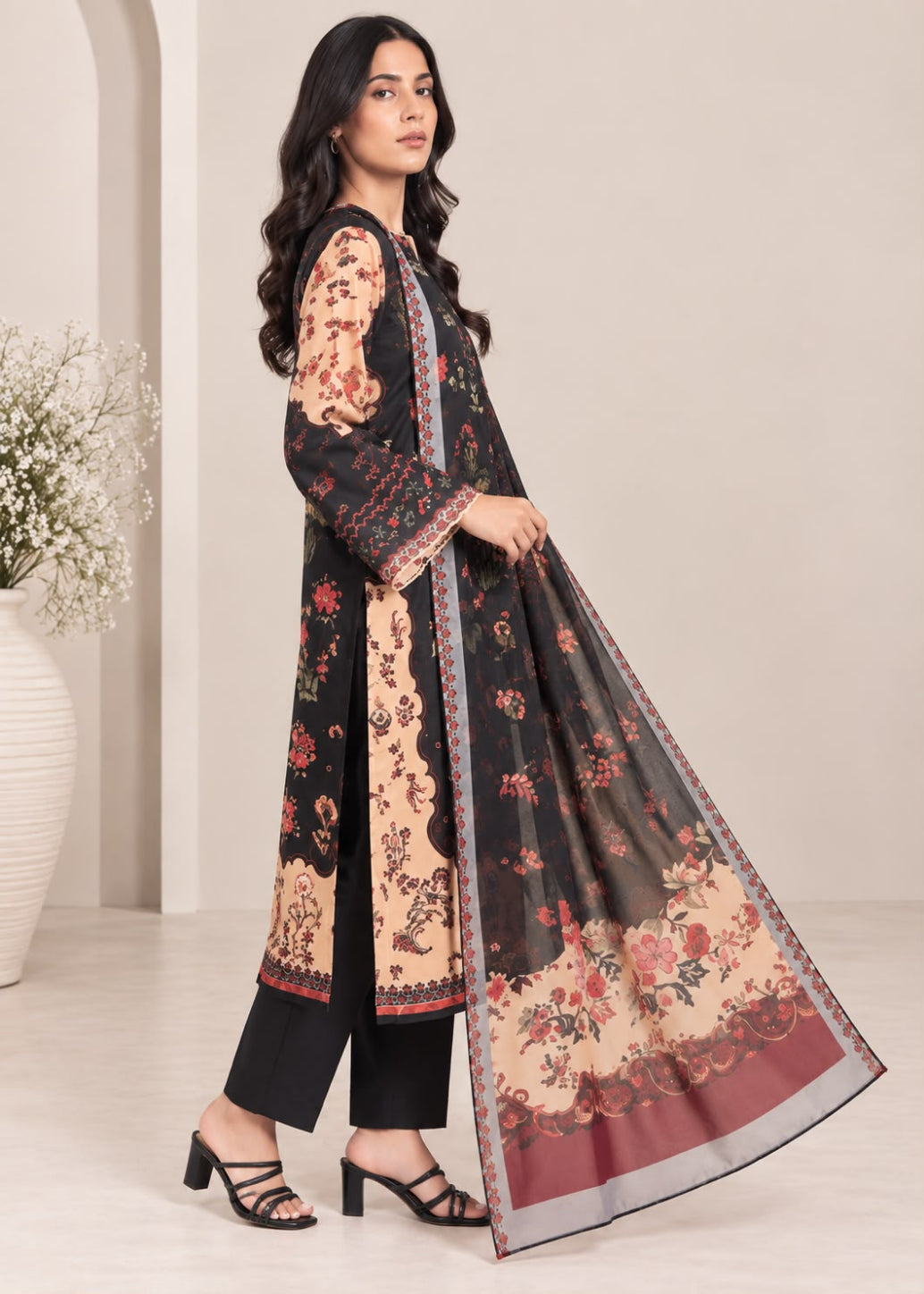 A woman models a traditional three-piece salwar kameez suit set. The set features a black kurta with beige floral patterns and red accents, black trousers, and a matching sheer dupatta with a beige border, floral print, and red bottom panel.