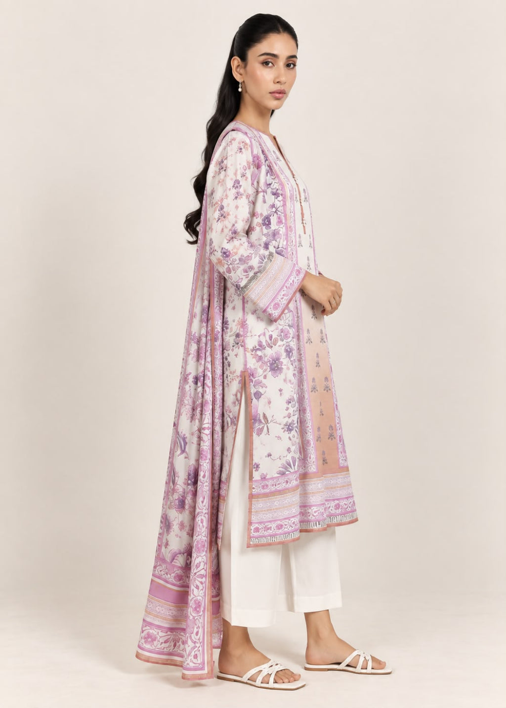 A woman modeling a traditional three-piece salwar kameez set. The outfit features a light pink and purple floral printed long kurta over white trousers, paired with a matching pink and purple dupatta.