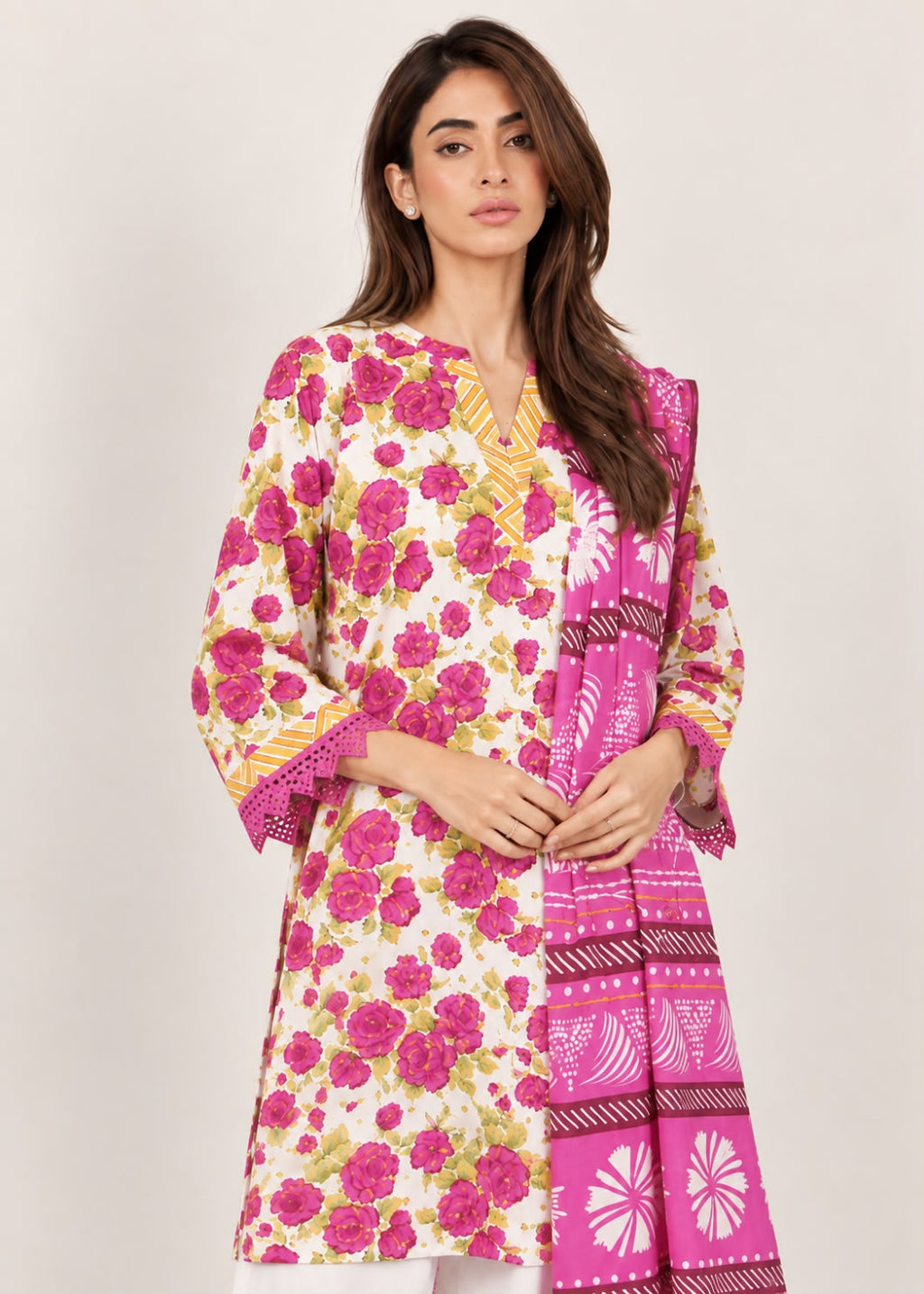 A woman modeling a three-piece traditional outfit featuring a cream-colored kurta with a vibrant pink and green floral print, a matching pink dupatta with geometric patterns, and a yellow embroidered border on the sleeves.