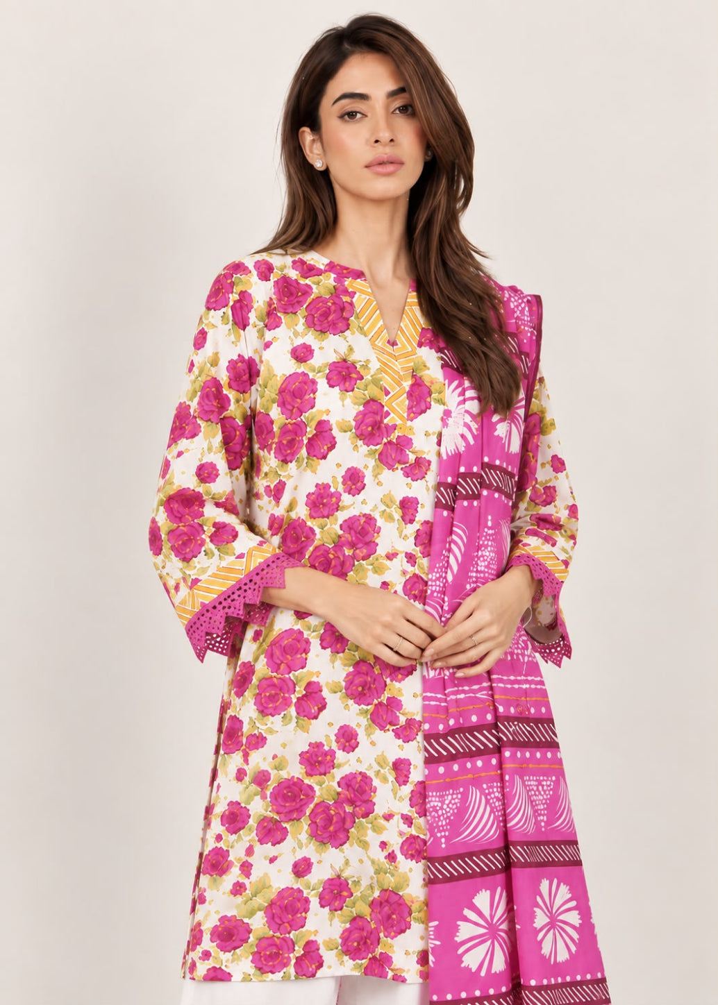 A woman modeling a three-piece traditional outfit featuring a cream-colored kurta with a vibrant pink and green floral print, a matching pink dupatta with geometric patterns, and a yellow embroidered border on the sleeves.