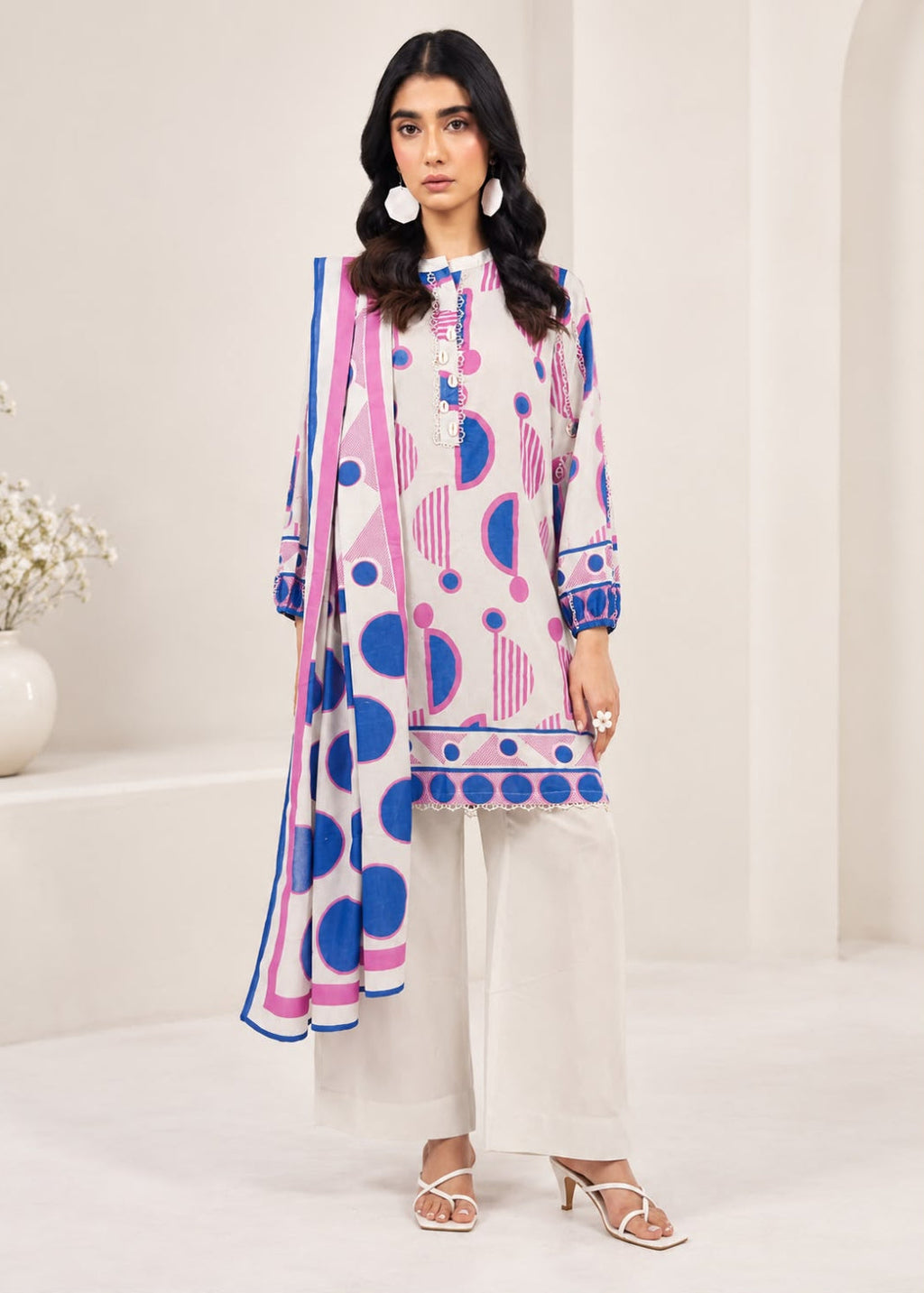 A woman modeling a three-piece printed salwar kameez suit in a bright, modern studio setting. The set features a white kurta and wide-leg trousers with pink and blue geometric patterns, paired with a matching dupatta.