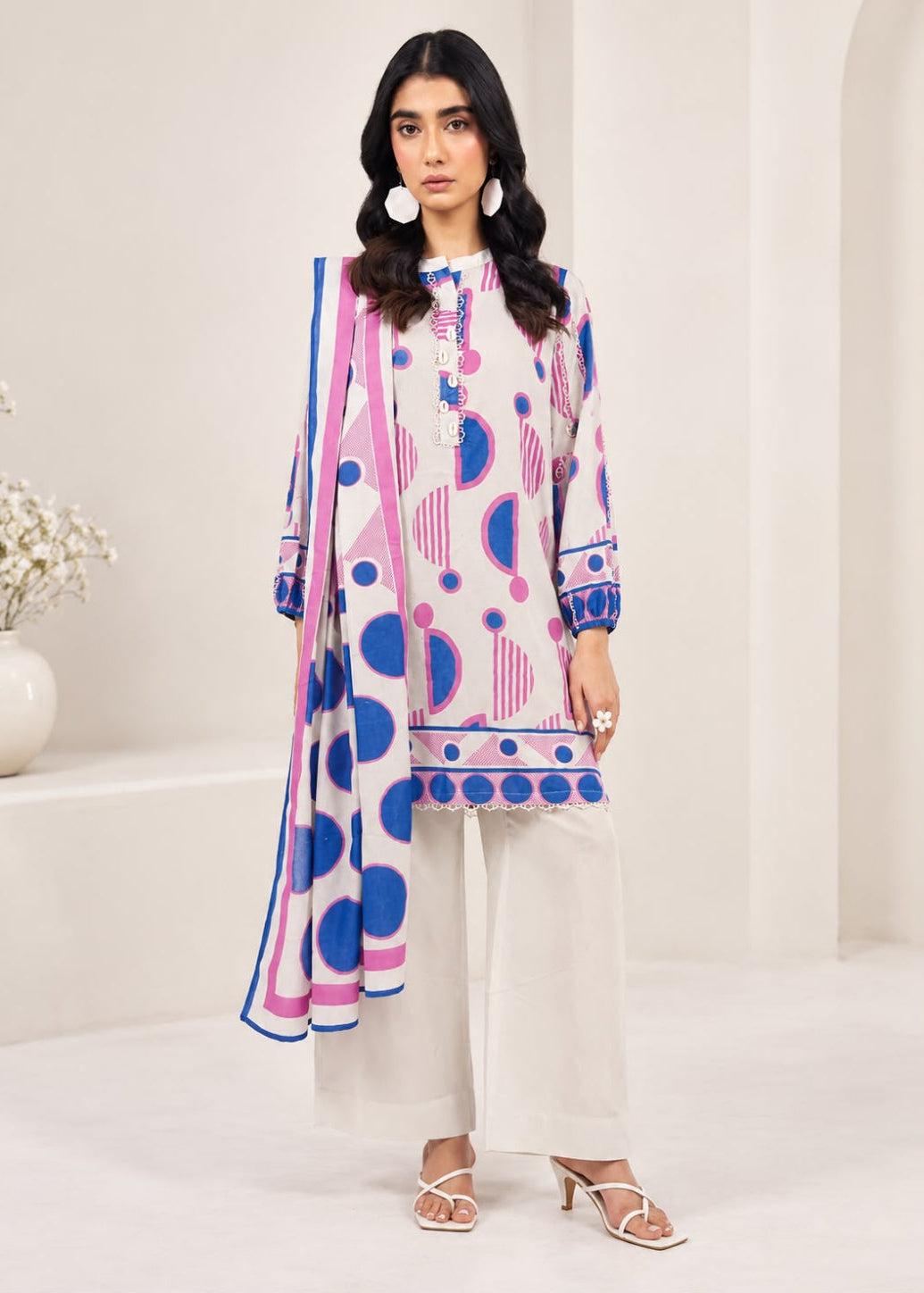 A woman modeling a three-piece printed salwar kameez suit in a bright, modern studio setting. The set features a white kurta and wide-leg trousers with pink and blue geometric patterns, paired with a matching dupatta.