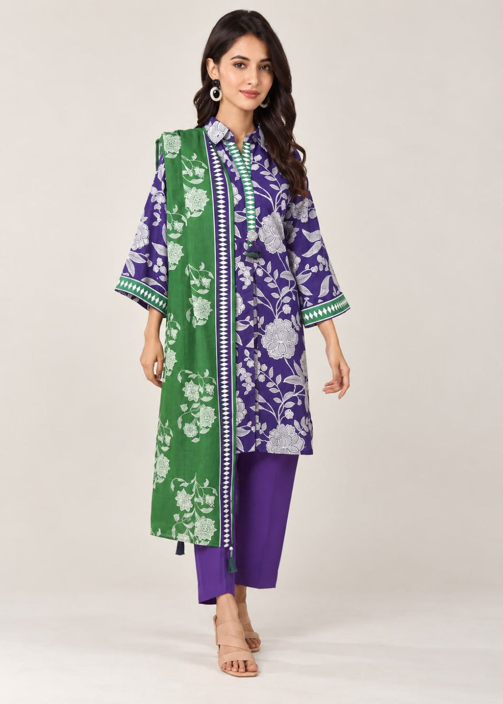 A woman models a traditional three-piece Indian outfit consisting of a purple and green floral printed kurta, solid purple pants, and a green dupatta with white floral patterns, set against a plain light beige studio background.
