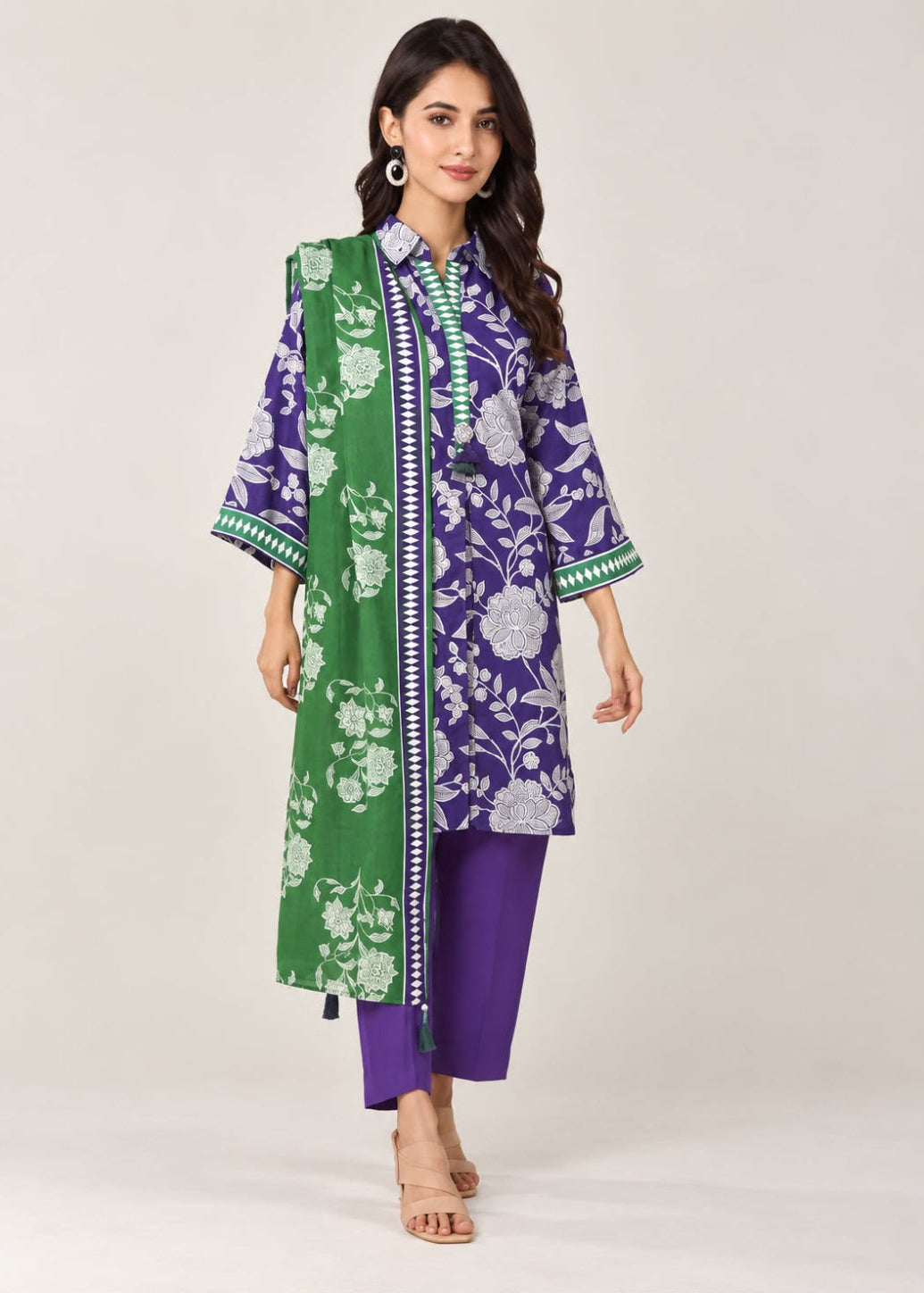 A woman models a traditional three-piece Indian outfit consisting of a purple and green floral printed kurta, solid purple pants, and a green dupatta with white floral patterns, set against a plain light beige studio background.