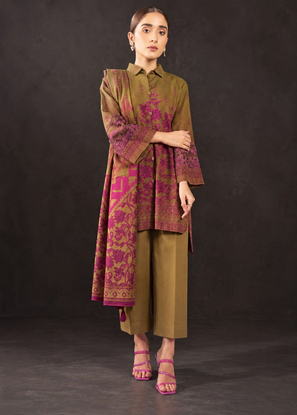 A woman models a traditional olive green three-piece outfit with magenta floral embroidery and block prints, consisting of a long-sleeve kurta, matching trousers, and a collared shirt, set against a dark studio backdrop.