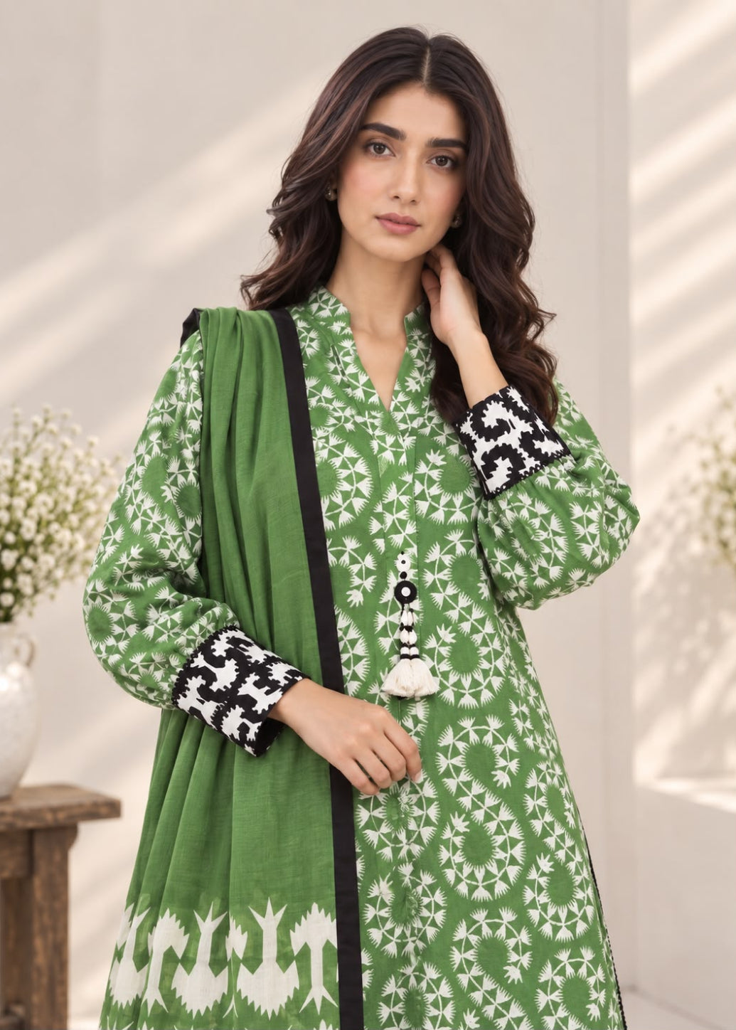 A woman modeling a green printed traditional outfit with a matching dupatta, featuring white geometric patterns, black-and-white embroidered cuffs, and a tassel detail, set against a softly lit indoor background.
