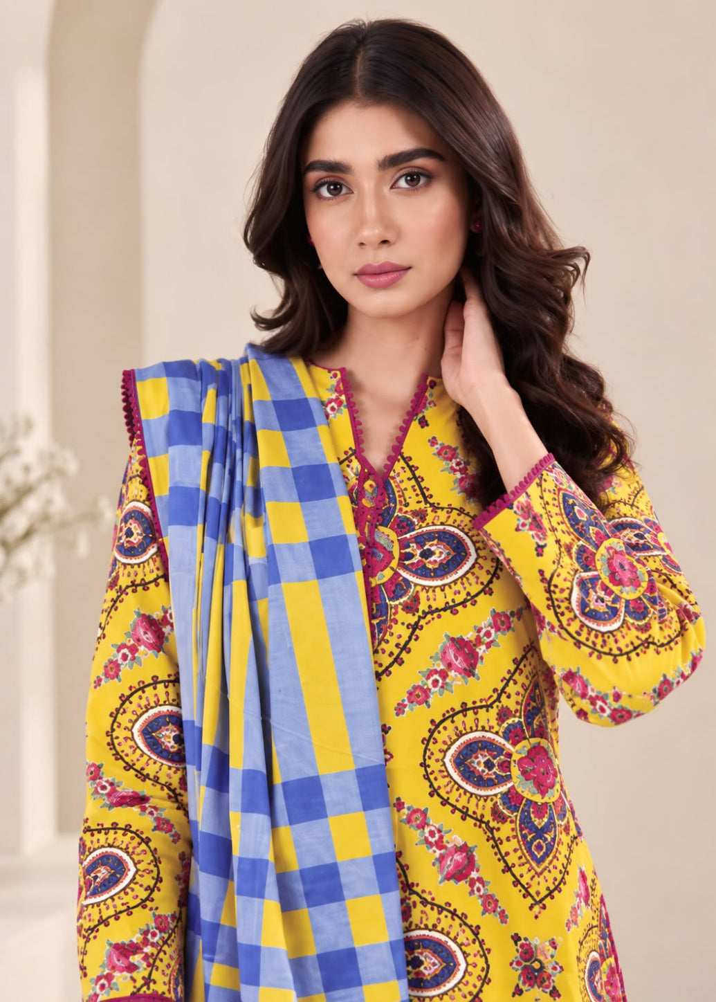 A woman modeling a yellow printed kurta with colorful paisley motifs and a blue and yellow checkered dupatta, set against a soft beige background.