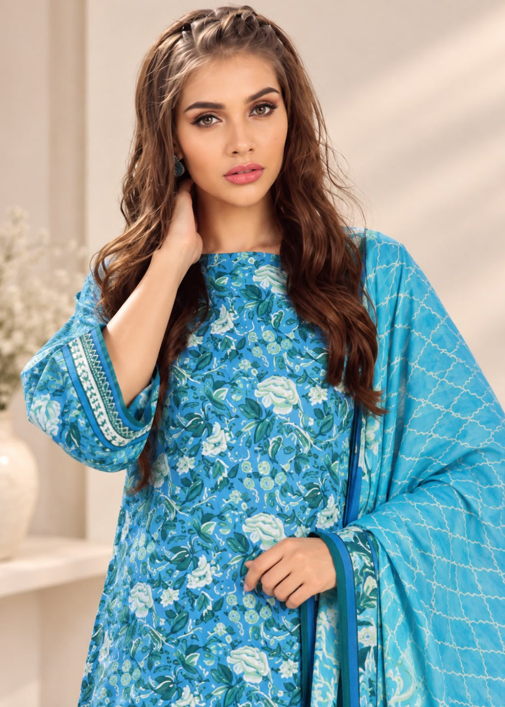 A woman modeling a bright blue floral salwar kameez suit with a matching printed dupatta, set against a softly lit beige indoor background.
