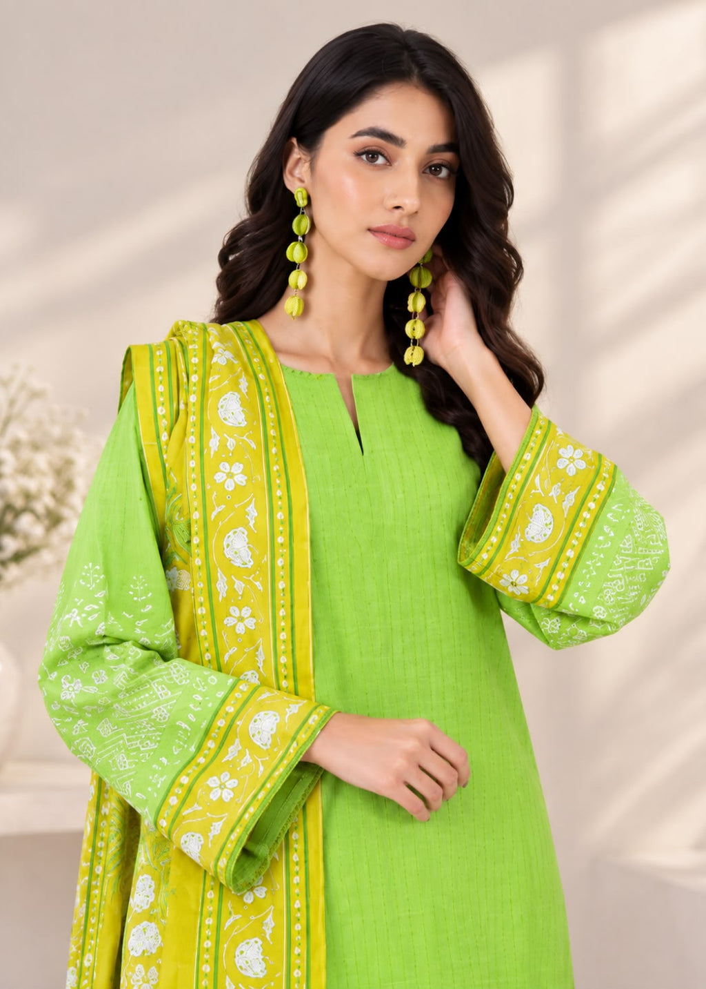 A woman modeling a bright lime green traditional outfit with matching printed dupatta and long yellow-green beaded earrings, posed against a softly lit neutral indoor background.