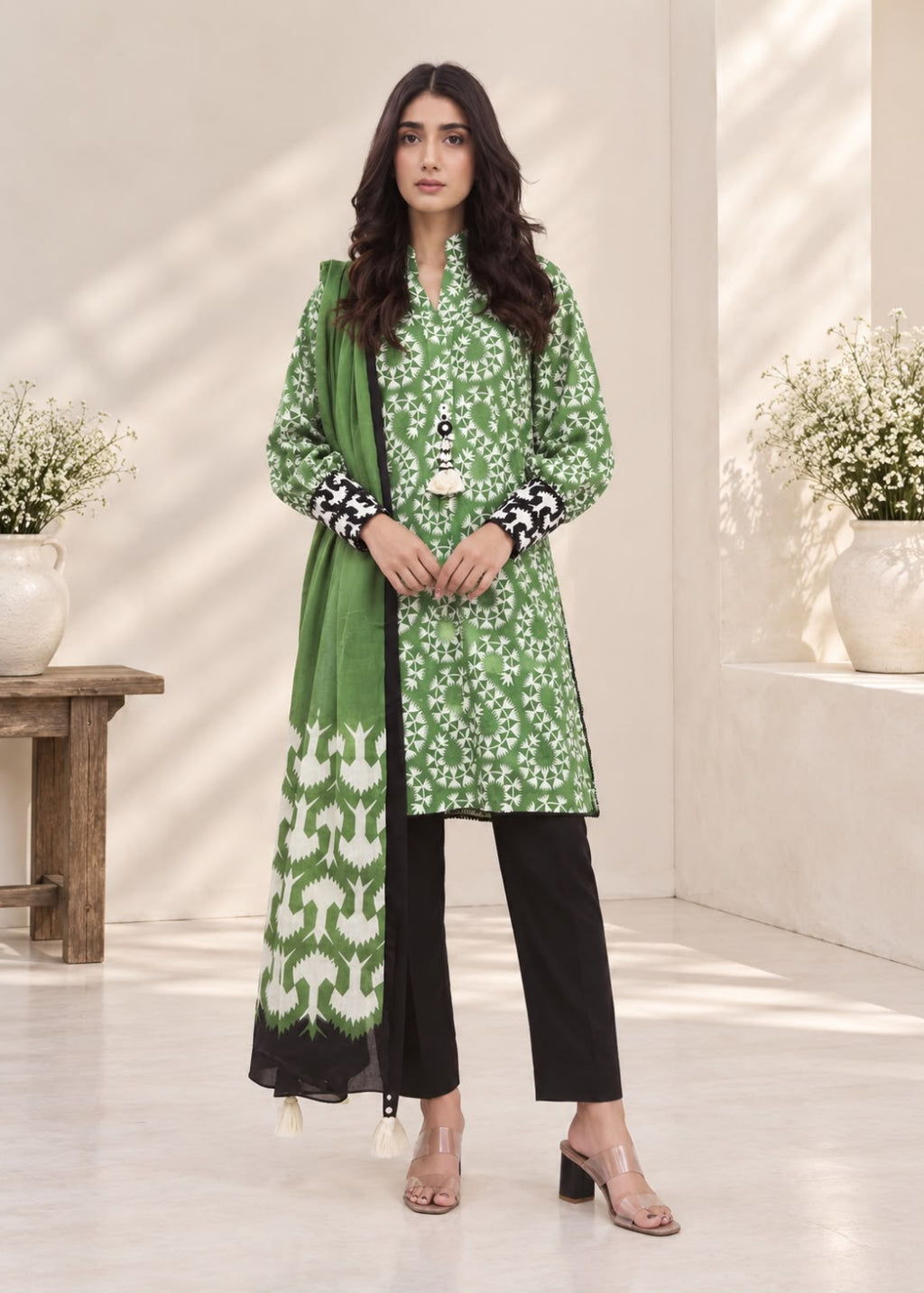 A woman models a three-piece green salwar kameez suit with white floral print, black pants, and a matching green dupatta with a white geometric border, set against a warm beige background with soft window shadows.
