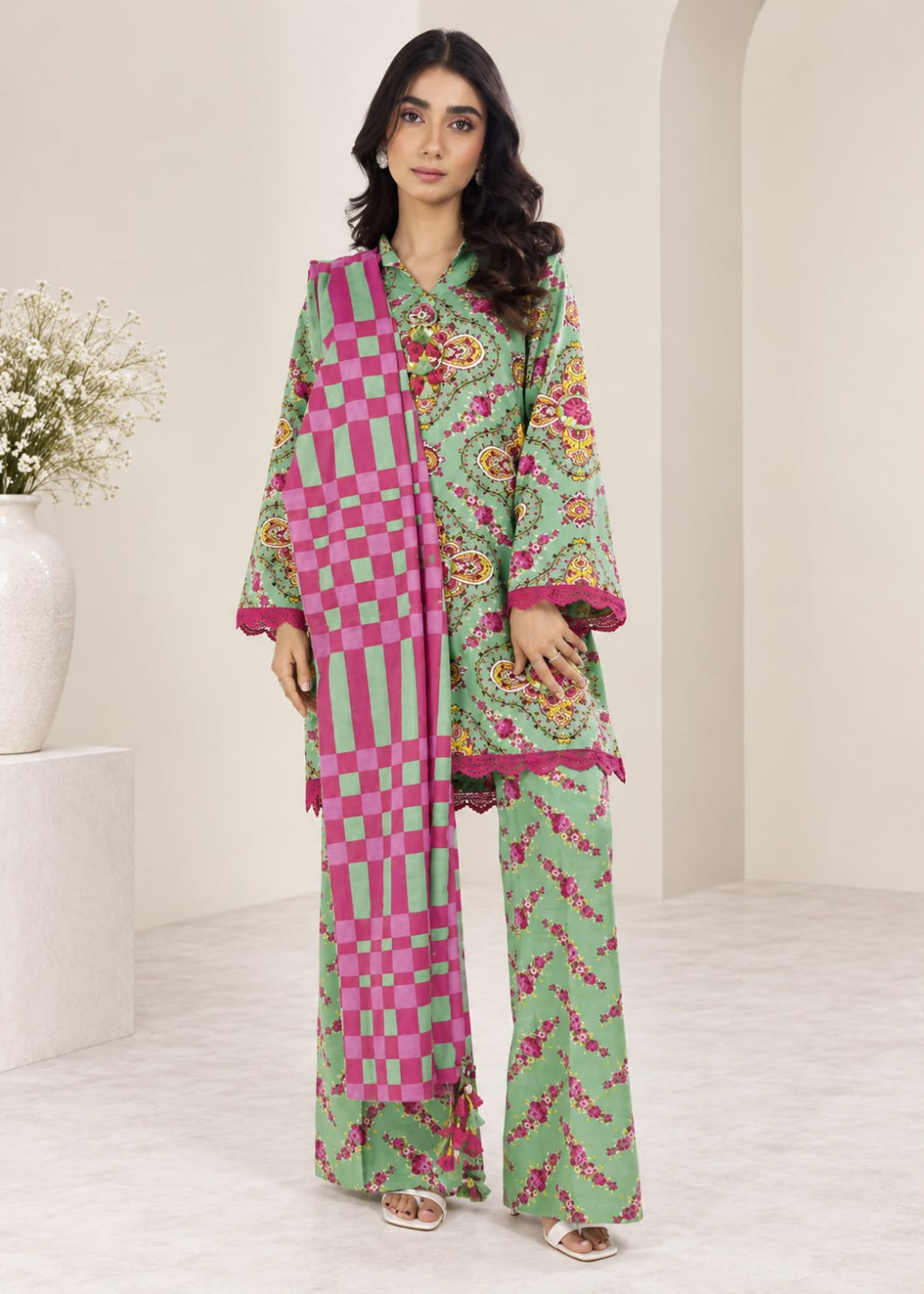 A woman modeling a three-piece green and pink traditional outfit with floral prints, checkered patterns, and wide-leg pants, standing in a minimalist beige room.