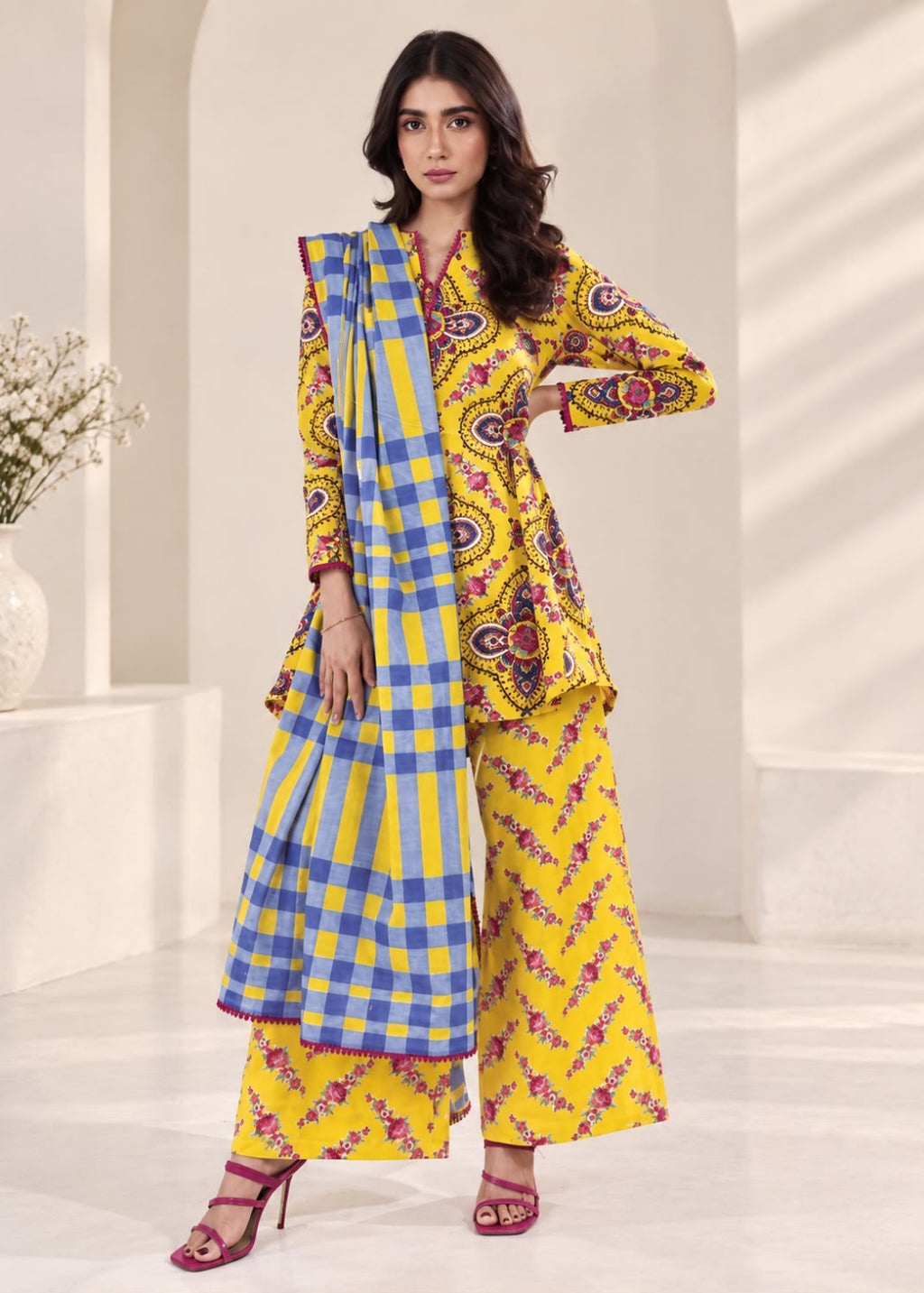A woman models a bright yellow three-piece ethnic suit with blue and purple paisley and checkered patterns, set against a clean, modern white architectural background.