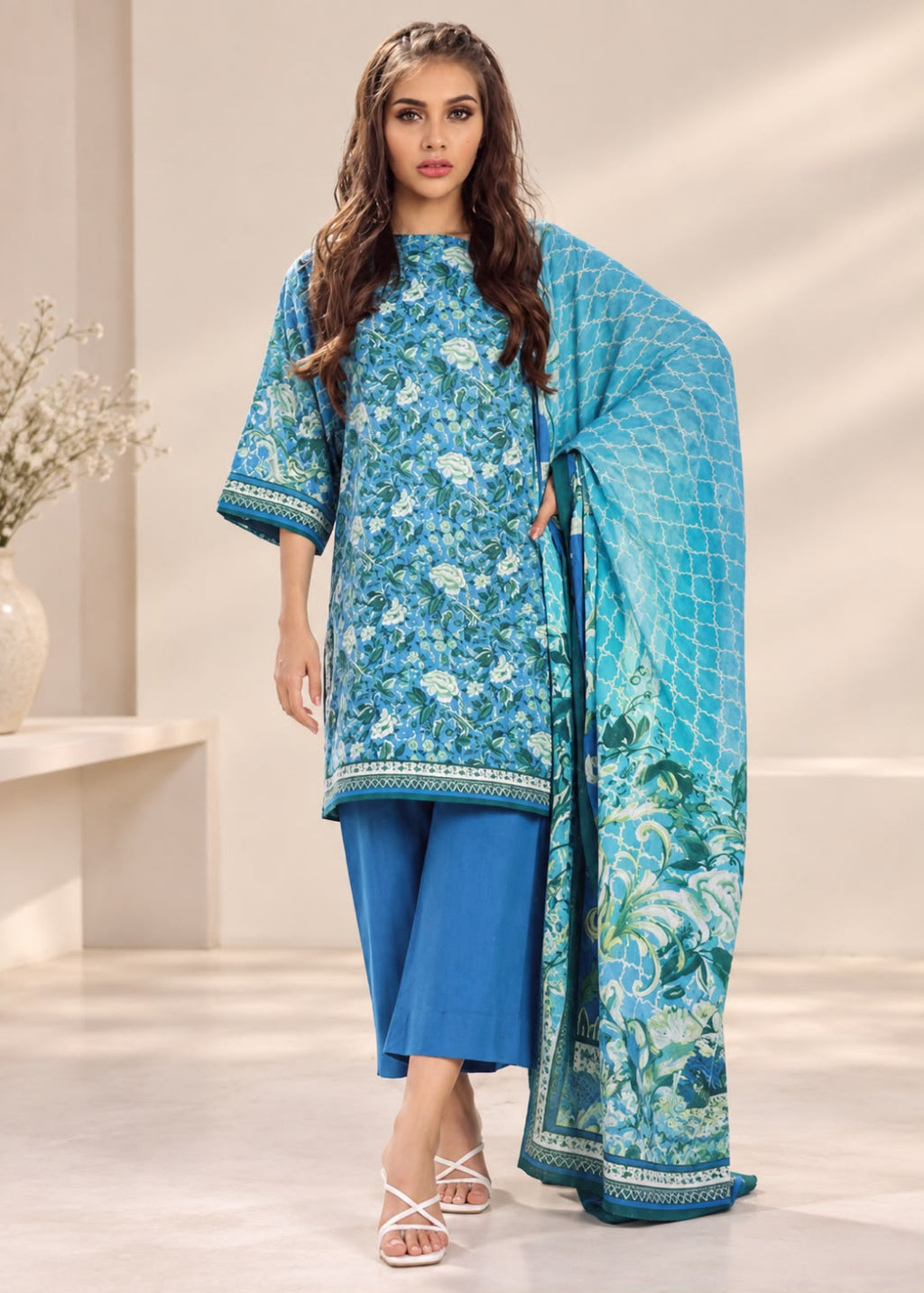 A woman models a traditional three-piece salwar kameez suit set in a bright, modern studio setting. The set features a light blue and white floral printed kameez kurta, solid blue cropped pants, and a matching light blue printed dupatta with a geometric pattern and a blue border.