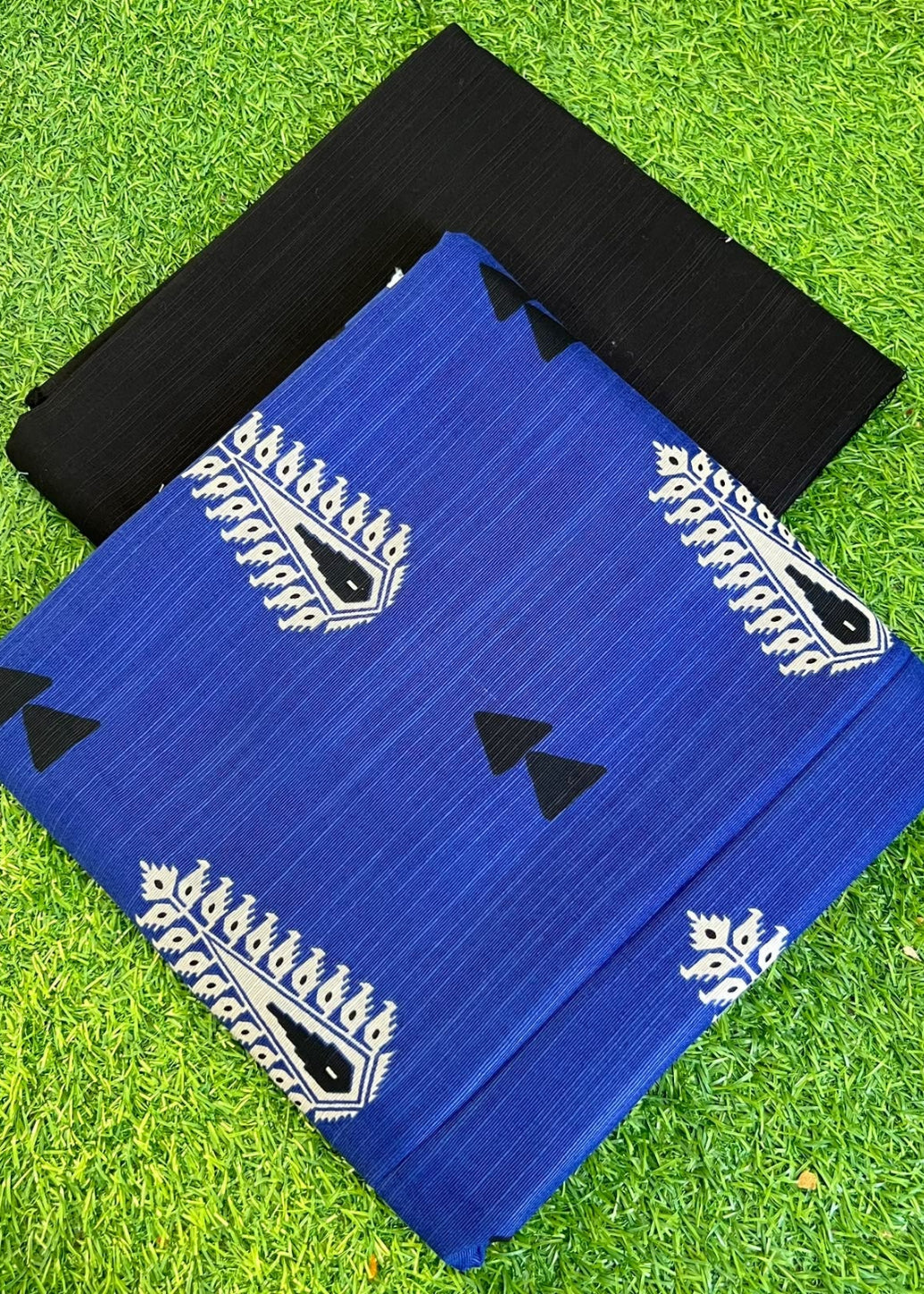 Two folded fabric pieces, one royal blue with white paisley and black triangle motifs, and one solid black, displayed on a green grass background.