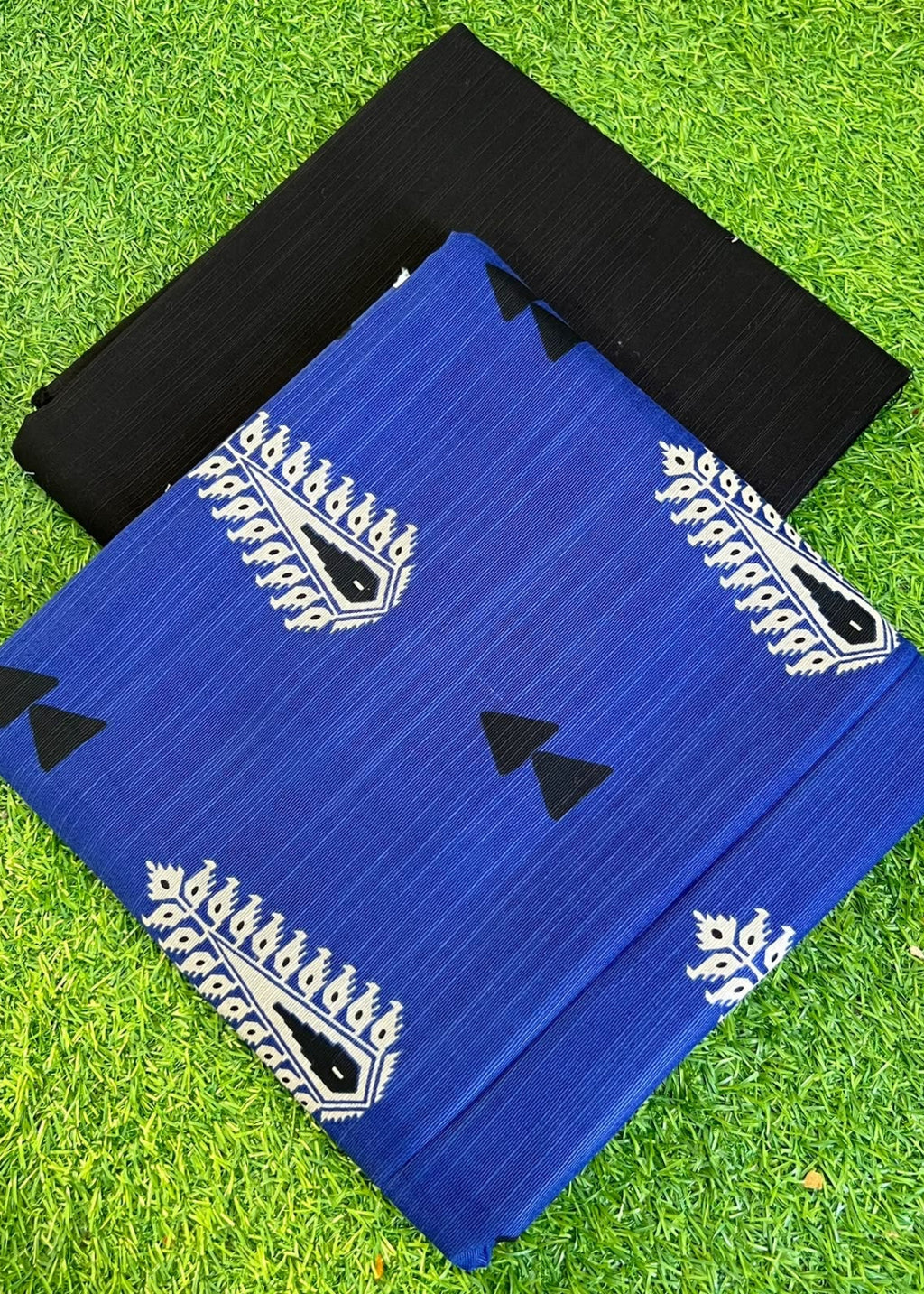 Two folded fabric pieces, one royal blue with white paisley and black triangle motifs, and one solid black, displayed on a green grass background.