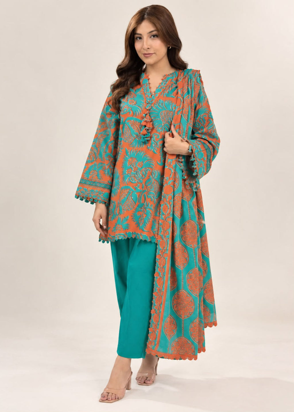 A woman modeling a traditional South Asian outfit featuring a teal and rust-orange patterned kurta with matching pants and a dupatta, set against a plain light beige studio background.