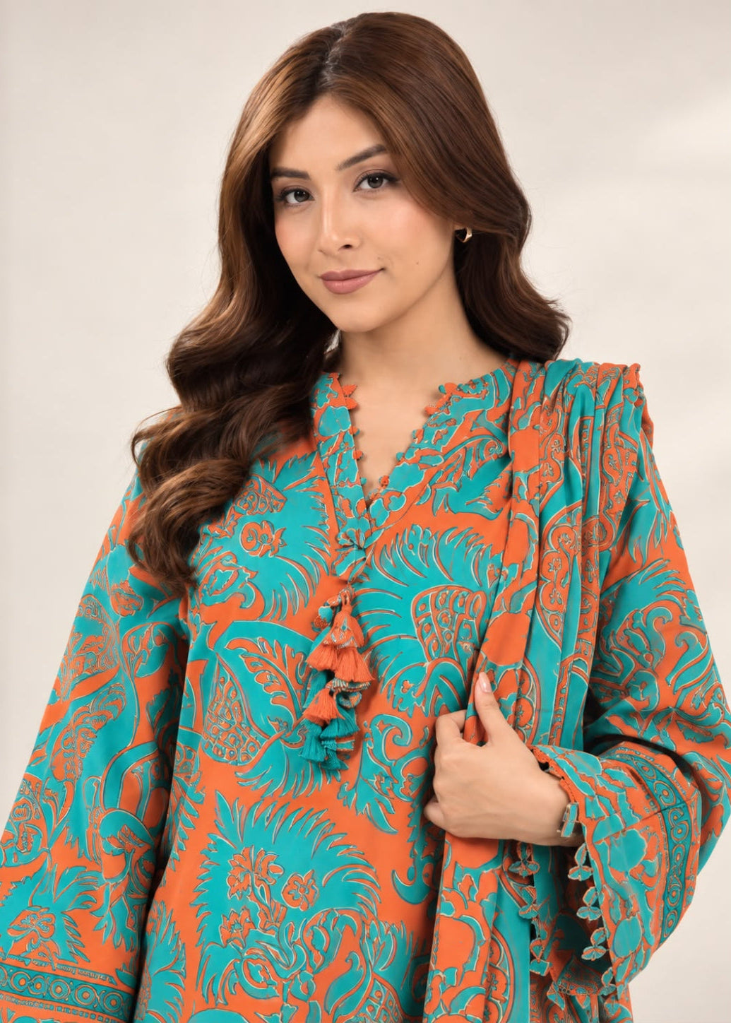 A woman wearing a vibrant teal and orange patterned tunic top with tassel ties, photographed against a neutral light beige background.