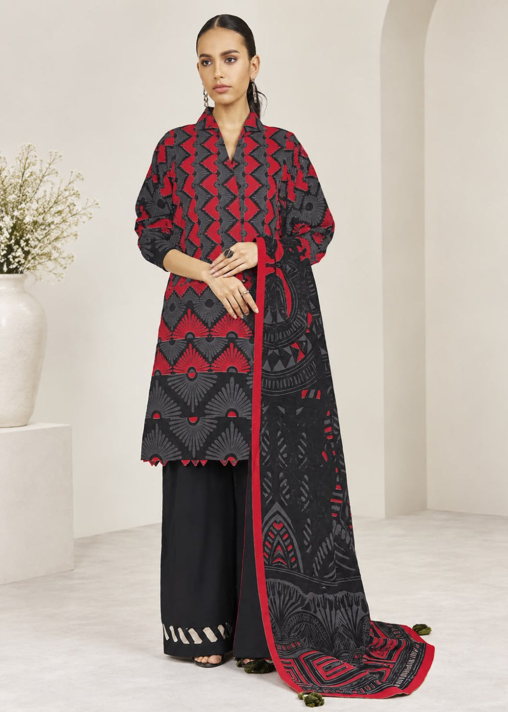 A woman models a three-piece traditional South Asian outfit consisting of a black and red patterned kurta, black wide-leg trousers, and a matching black dupatta with red borders, set against a minimalist cream-colored interior background.