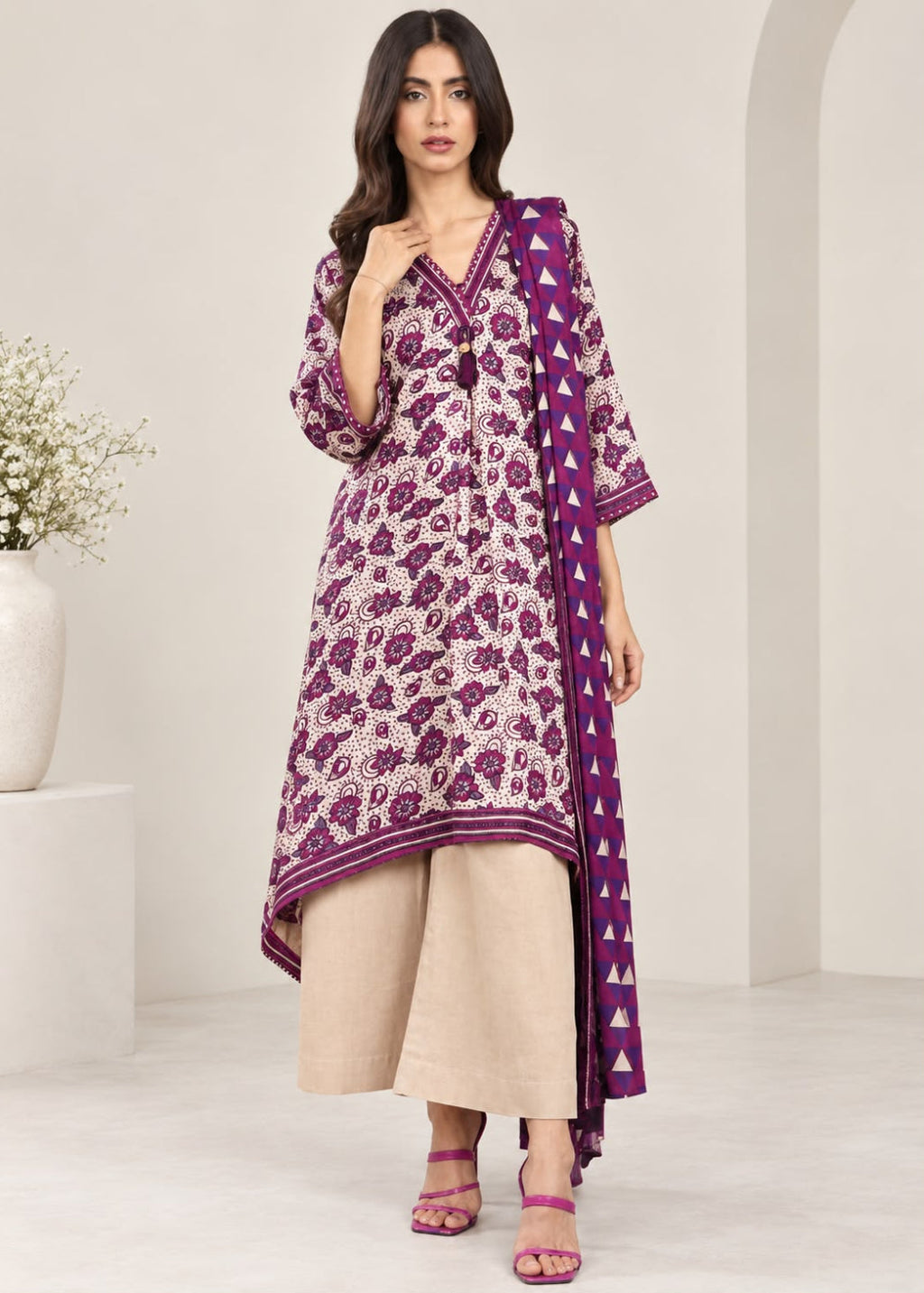 A woman models a traditional three-piece Indian salwar kameez set. The set features a purple and beige floral print kurta with a matching printed dupatta, paired with wide-leg beige trousers and pink sandals, set against a minimalist cream-colored studio background.