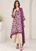 A woman models a traditional three-piece Indian salwar kameez set. The set features a purple and beige floral print kurta with a matching printed dupatta, paired with wide-leg beige trousers and pink sandals, set against a minimalist cream-colored studio background.
