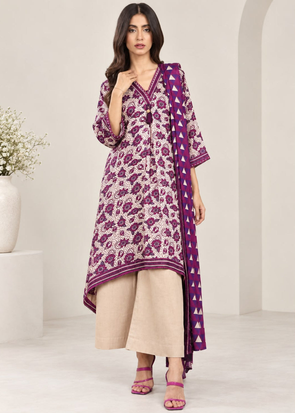 A woman models a traditional three-piece Indian salwar kameez set. The set features a purple and beige floral print kurta with a matching printed dupatta, paired with wide-leg beige trousers and pink sandals, set against a minimalist cream-colored studio background.