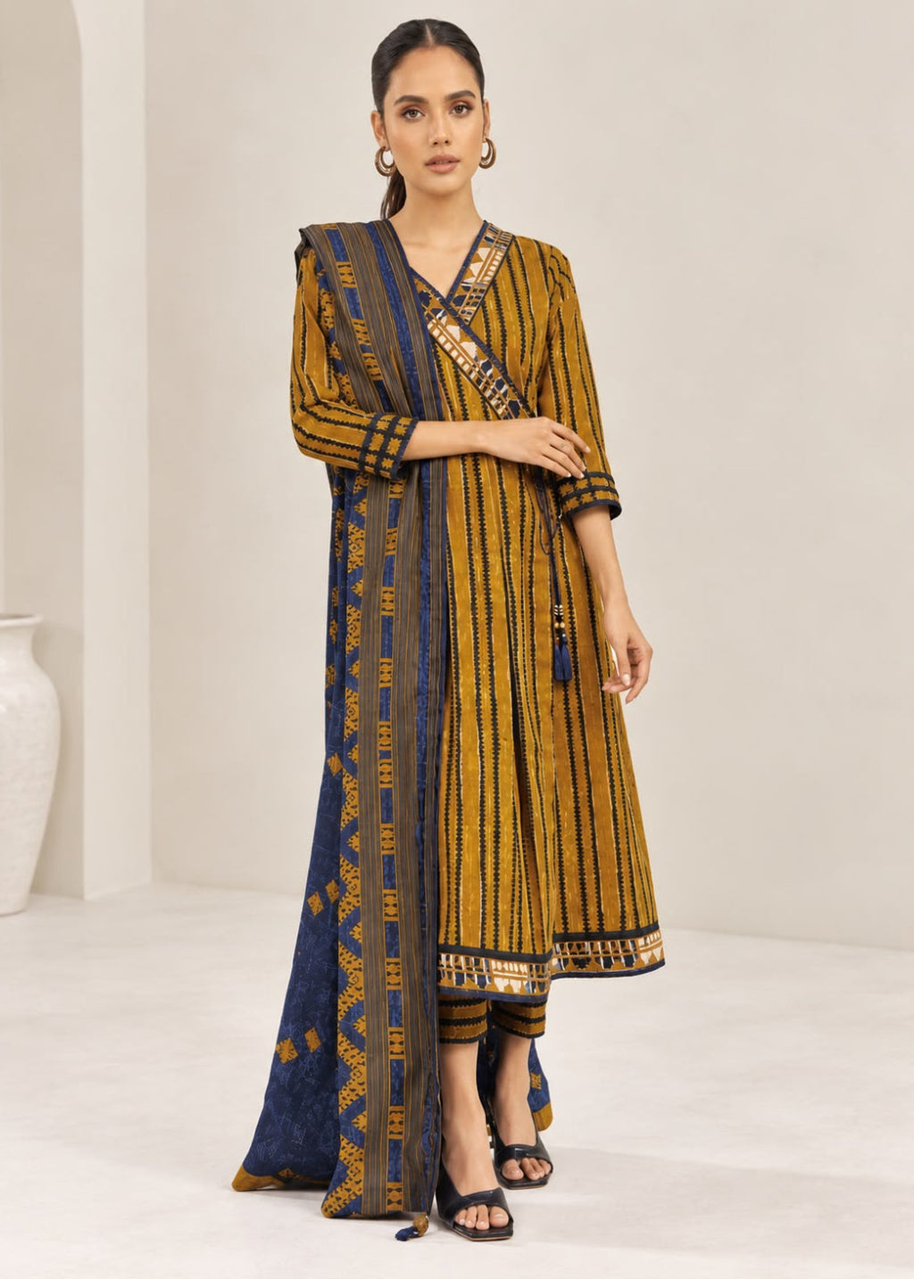 A woman models a traditional Indian outfit consisting of a mustard yellow and navy blue striped kurta, matching striped pants, and a navy blue dupatta with gold patterns, set against a minimalist beige studio background.