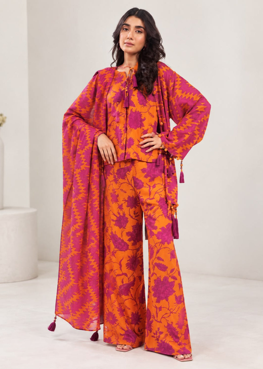 A woman modeling a vibrant orange and magenta floral print three-piece traditional outfit with a matching draped dupatta, set against a clean, minimalist studio background.