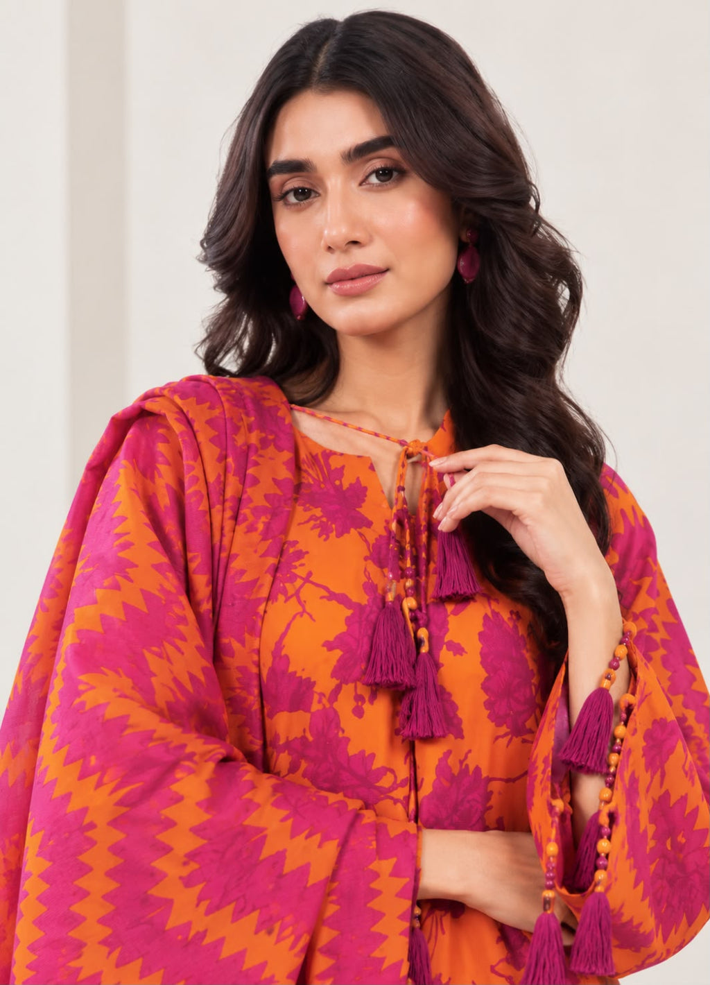 A woman wearing a bright orange and magenta patterned tunic with matching tassel necklaces, photographed against a clean light beige background.