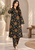 A woman models a black three-piece salwar kameez suit with colorful floral prints, set against a warm, neutral-toned architectural background with a large vase and wooden stool.