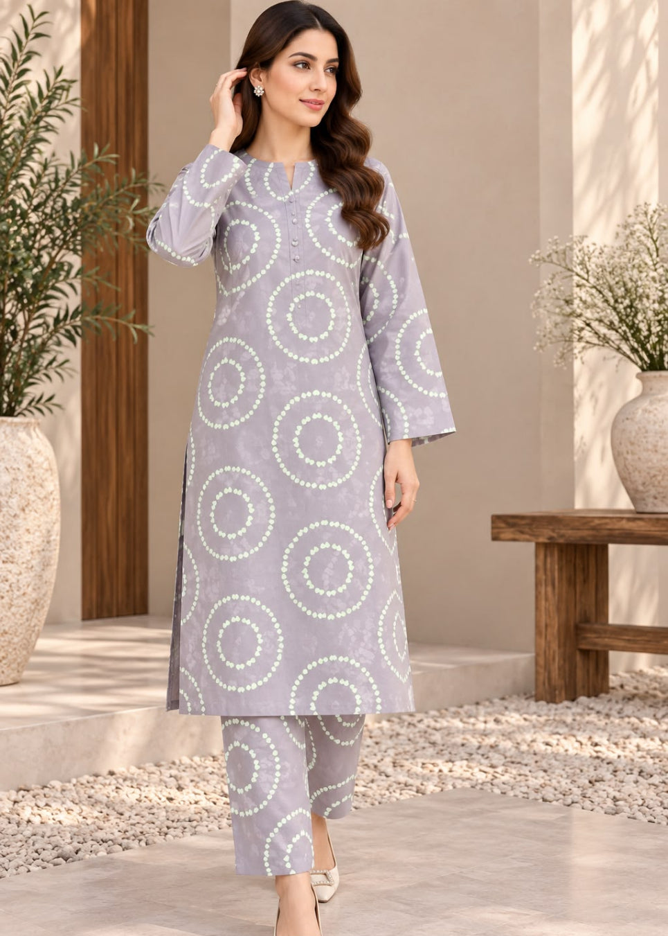 A woman models a light lavender-gray three-piece kurta set with white circular dot patterns, standing in a styled outdoor patio setting with plants and a wooden bench.