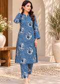 A woman models a blue floral printed kurta and trouser set in an outdoor patio setting with beige walls, potted plants, and a wooden bench.