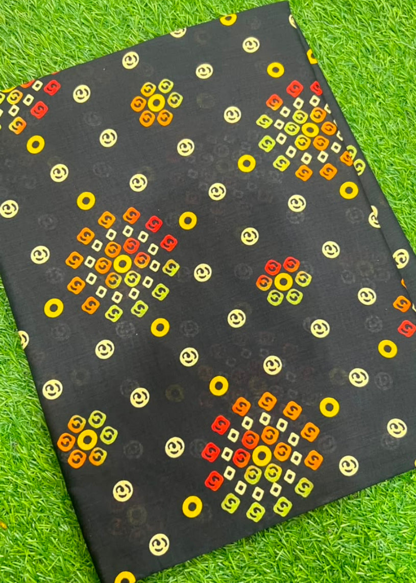 A black fabric cloth with a colorful geometric pattern of yellow, orange, and red smiley faces, laid flat on a green grass background.