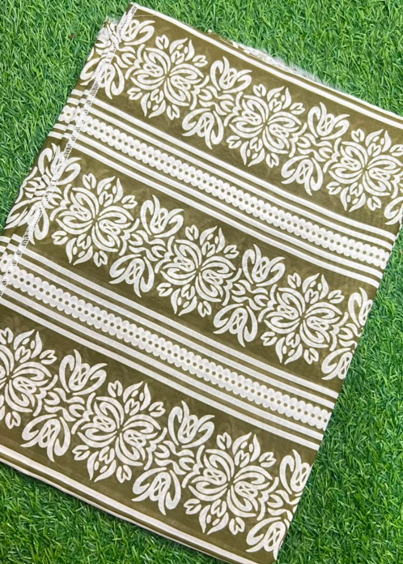 A rectangular fabric item, likely a tablecloth or napkin, featuring a white floral and geometric block print on an olive green background, laid flat on green grass.