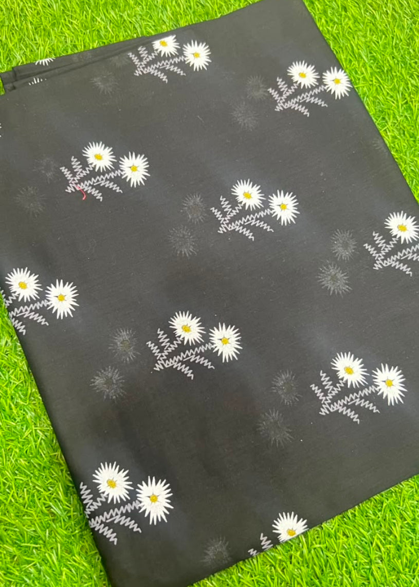 A black fabric item, likely a scarf or sari, featuring a repeating pattern of white daisies with yellow centers and gray leafy stems, laid flat on a bright green grass background.